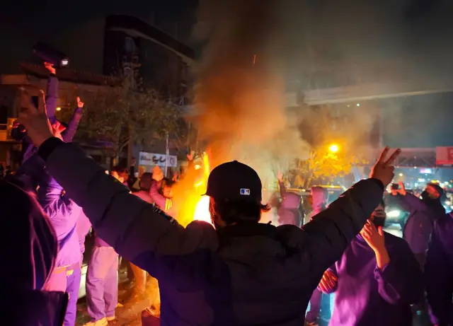 معترضی از پشت دست‌هایش را باز کرده است با نشان پیروزی روبه روی آتش. معترضان دیگر هم دور آتش ایستاده‌اند. 