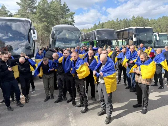 Велика група людей стоїть на відкритому майданчику перед кількома автобусами. Багато з них загорнуті в синьо-жовті українські прапори. На тлі — дерева та ліс, сонячний день. 