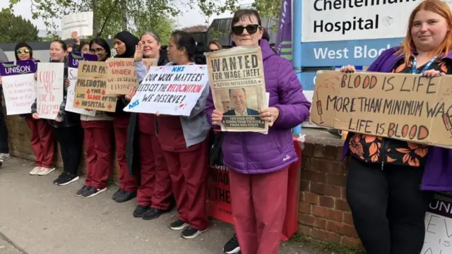 A group of women in red hospital scrubs and coats stands in front of a low brick wall on a picket line holding placards protesting at their level of pay.
