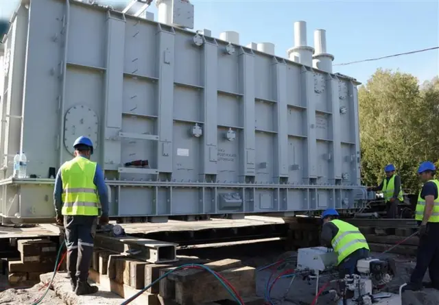 На малюнку - трансформатор білого кольору, поруч із ним чотири працівники в жовтих желетах та касках налаштовують його роботу