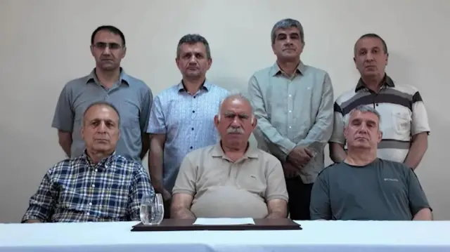Öcalan mira a la cámara mientras está sentado frente a una mesa con un mantel azul claro, un individual negro, un vaso de agua y una hoja de papel. Lo rodea un grupo de 6 hombres más quienes también miran a la cámara.