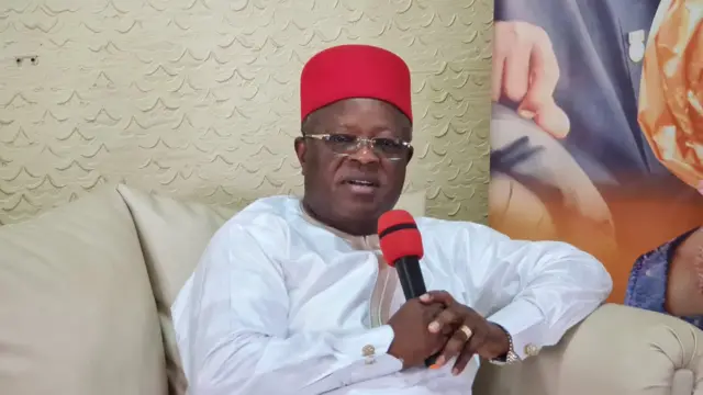 David Umahi zaune kan kujeri mai launin ruwan riƙe da abin magana mai launin baki da jan kai, sanye da fararen tufafai da jar dara