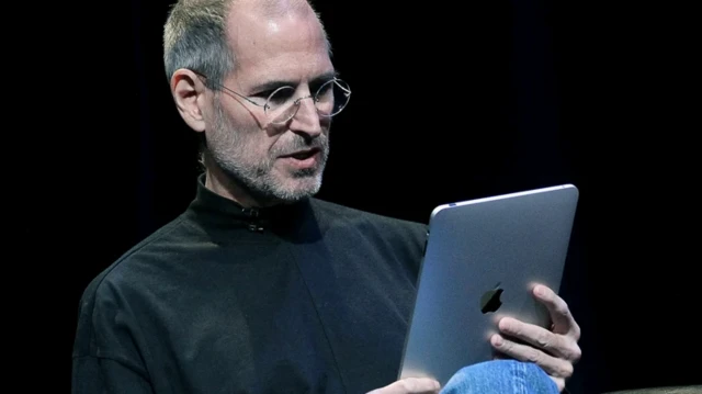 استیو جابز که نشسته، یک آیپد در دست دارد و به آن نگاه میکند