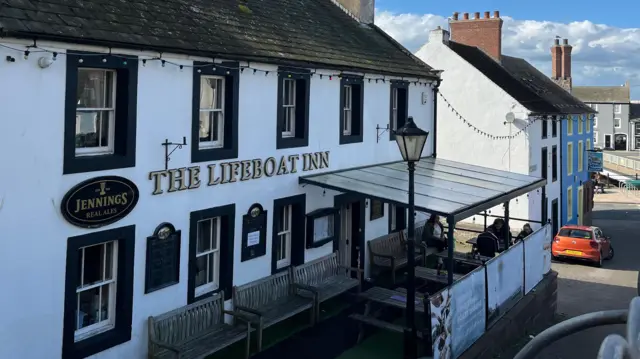 El Lifeboat Inn de Maryport. El edificio blanco de dos plantas tiene detalles negros alrededor de las ventanas y la puerta. El nombre está en la fachada en letras doradas. Una zona elevada en la parte delantera del pub está cubierta por una marquesina con estructura metálica y lona transparente.