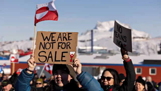 Protesters gather outside the US consulate during a demonstration in Nuuk, Greenland, on March 15, 2025. In the foreground, a woman wearing a blue-and-black puffer jacket holds a banner reading “We are not for sale”. Behind her, another woman in a black puffer jacket and sunglasses holds a sign that reads “No means no” while several Greenland flags are visible in the crowd.