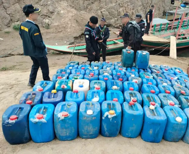 မြစ်ကမ်းဘေးရပ်ထားတဲ့ လှေ ၂ စီးနောက်ခံနဲ့ ကမ်းပေါ်ချထားတဲ့ ဂါလံပုံး အပြာရောင်တွေ၊ ဘေးမှာရပ်နေကြတဲ့ ထိုင်းအကောက်ခွန်အရာရှိတွေ 