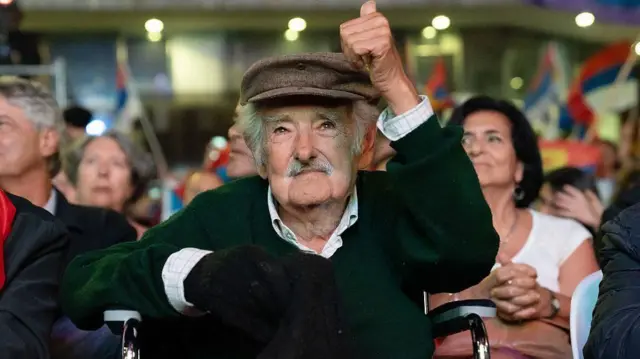 Uwahoze ari Perezida wa Uruguay José Mujica azamuye igikumwe, ubwo yari yicaye mu mbaga y'abantu, mu gikorwa cyabaye mbere mu mwaka wa 2025.