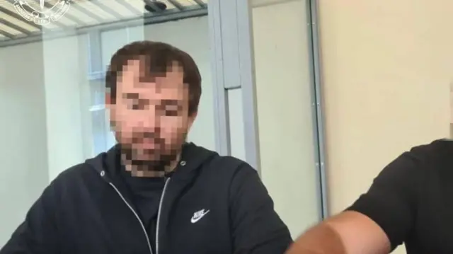 Чоловік у чорній кофті у залі суду. Його обличчя заблюрене.
