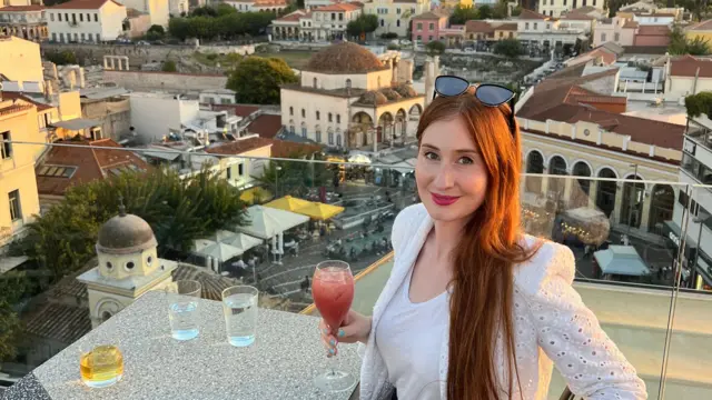 Amber siedzi przy stoliku w ogródku restauracyjnym w Atenach. W tle widoczny jest Akropol.