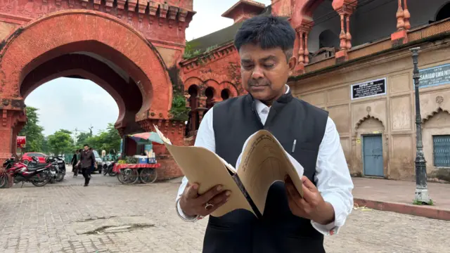 शाहिद अली ख़ान हाथों में कुछ पेपर लिए खड़े हुए
