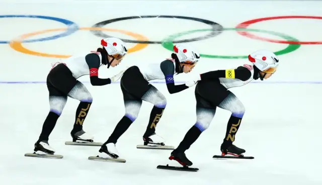 3人が縦に並んで滑っているのを横からとらえた写真。後方の氷上に五輪マークが描かれている