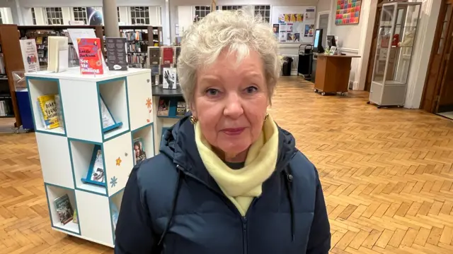 Una mujer de cabello blanco mira directamente a la cámara. Lleva pendientes, una bufanda amarilla y una chaqueta azul marino. Detrás de ella hay estanterías de biblioteca y un suelo de madera con dibujos.