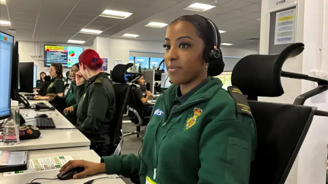Ugbad Yousuf, coordinadora de llamadas de emergencia, se encuentra en la sala de control del Servicio de Ambulancias de Londres. Viste un uniforme verde y tiene el pelo negro, sentada en un escritorio con su portátil y auriculares.
