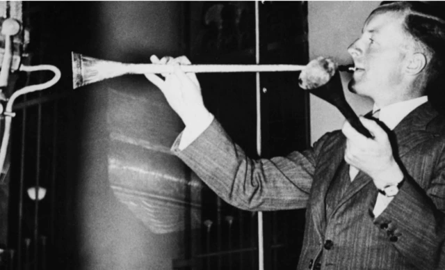 Siyah beyaz bir fotoğrafta, takım elbiseli bir kişi elinde trompet benzeri bir nefesli çalgıya üflüyor