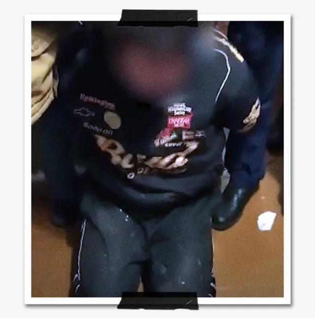 Фото Дмитрия Копылова по время задержания — темноволосый мужчина в спортивной чёрной куртке с логотипом Bud и спортивных штанах стоит на коленях в помещении с коричневым полом, его руки за спиной в наручниках. 