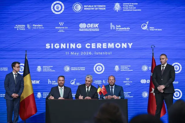Belçikalı ve Türk yetkililer bir kürsüdeler ve imza töreni yapıyorlar. Toplam beş erkek var.