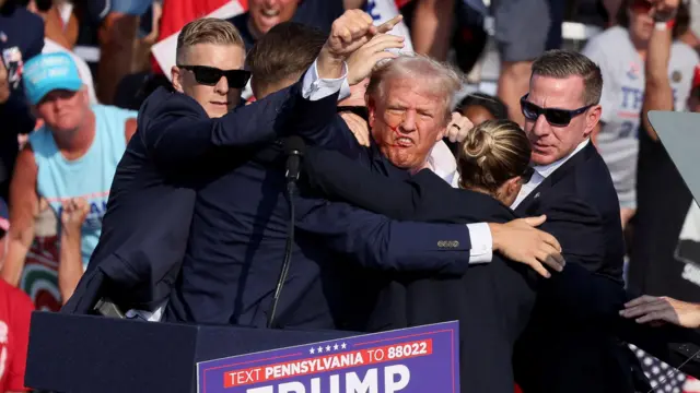 Trump, ainda no palco, rodeadoroleta roubaagentes da segurança, com a mãoroleta roubapunho para o alto e o lado direito do rosto com sangue. 