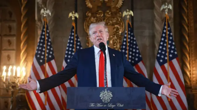 Donald Trump está vestindo um terno azul marinho sobre camisa branca e gravata vermelha. Ele está com os braços abertosjoguinho que ganha dinheiroum púlpito. Atrás dele, quatro bandeiras dos EUA.