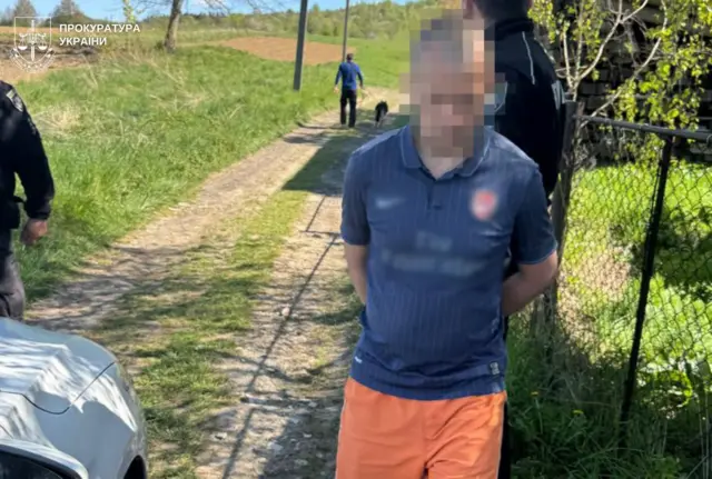 Чоловік із розмитим обличчям стоїть на ґрунтовій дорозі біля паркану в сільській місцевості. Він одягнений у блакитну футбольну футболку та помаранчеві шорти, руки тримає за спиною (ймовірно, у кайданках). Поруч видно автомобіль, поліцейських і чоловіка з собакою на задньому плані. 