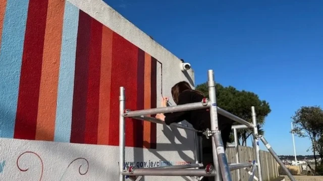 La imagen muestra una pared con rayas azules, rojo oscuro y naranja. Hay una persona de pie sobre un andamio pintando una raya rojo oscuro al final de una hilera de rayas de otros colores. Al fondo, hay árboles y el cielo es azul.