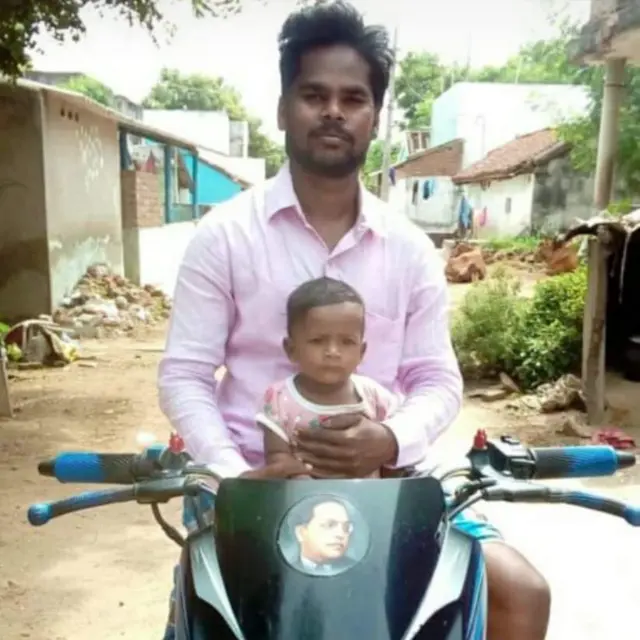 Nirmal, jeune enfant, assis sur une mobylette et câliné par son père Balaji.