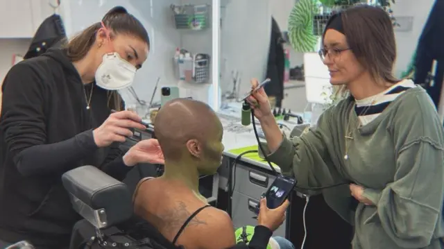 Cynthia Erivo está sentada en una silla de maquillaje negra en el tráiler de una película. Mira su teléfono mientras la maquillan y no mira a la cámara. Olivia Jerrard, a la izquierda, usa una brocha de maquillaje cerca de la oreja de Erivo, viste de negro y lleva una mascarilla blanca. A la derecha de Erivo está Alice Jones, quien usa un aerógrafo para pintar a Erivo de verde.