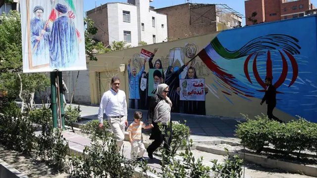 Двое взрослых и маленький ребенок идут по улице в Тегеране, украшенной проправительственными граффити