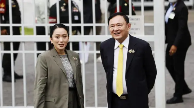 裁判所の白い格子の門の前に立つペートンタン氏とタクシン氏。ペートンタン氏はグレージュのスーツを着ている。背後には警備員や裁判所職員の姿が見える