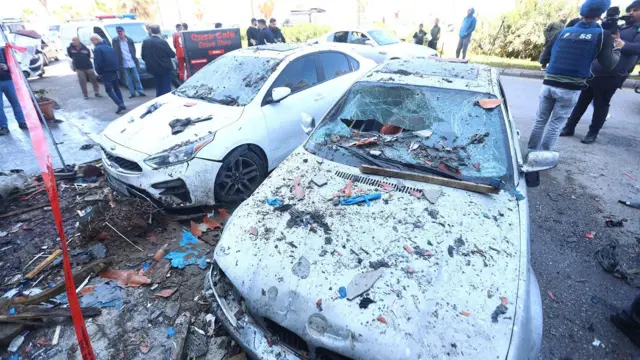 En la imagen aparecen dos automóviles dañados y cubiertos de escombros, con multitudes de personas en la calle de fondo y ruinas tras un ataque con misiles israelíes en la carretera costera de Sidón este miércoles.