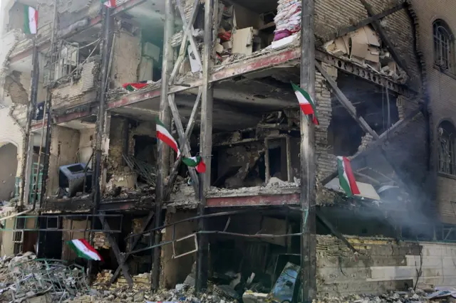 Edificio de varios pisos destruido. En medio de los escombros se ven banderas de Irán 