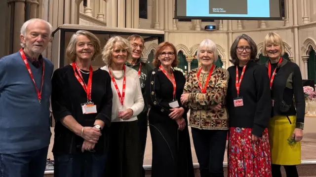 Un grupo de personas está de pie junto a otras en una iglesia. Llevan cordones rojos y sonríen a la cámara.