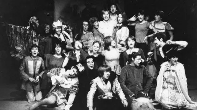 Una imagen en blanco y negro de varios niños participando en una actividad teatral. Algunos están sentados, otros de pie y otros arrodillados. Hacen muecas.