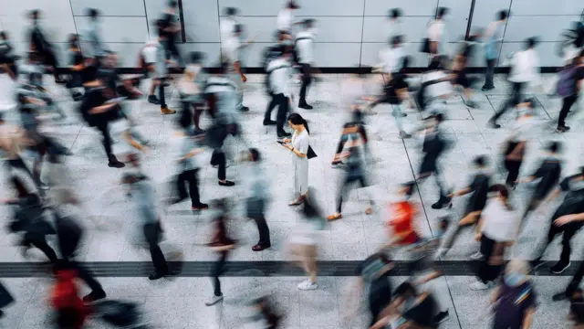 Egy nő áll a járda közepén, kezében lévő mobiljára koncentrál, miközben az emberek elmosódva rohannak el mellette.