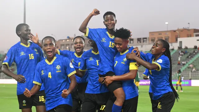 Un groupe de sept footballeuses tanzaniennes vêtues de maillots bleus à bordures jaunes et de shorts noirs célèbrent un but, l'une d'entre elles levant le poing en l'air tandis qu'elle est portée par une coéquipière.