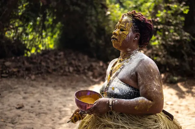 Une femme avec une tenue traditionnelle faite de paille autour du bassin, de la poudre blanche et de l'huile rouge sur le corps. Elle tient une assiette en plastique dans sa main gauche et semble être en transe.