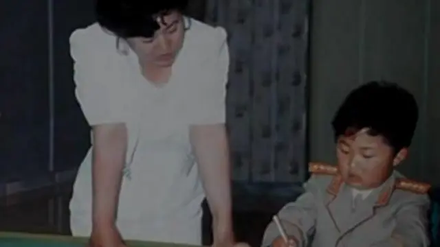 Ko Yong-hui, em um vestido branco, à esquerda. Ao lado dela, sentado, está Kim Jong-un, ainda criança, vestindo uniforme militar e escrevendo com uma caneta. 