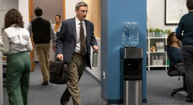 Un hombre con traje y corbata recorre una oficina donde otras personas van y vienen. 