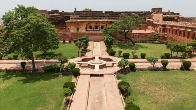 Espaço dividido em quatro jardins verdes, cruzados por quatro canais que chegam ao centro, onde há uma fonte. Existem também árvores e uma casa ao fundo.