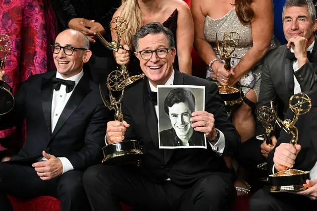 Stephen Colbert mira al frente, vestido de etiqueta sostiene el Emmy en su mano derecha y una foto suya de cuando era joven en la mano izquierda. A su alrededor hay otros ganadores con sus estatuillas en la mano.
