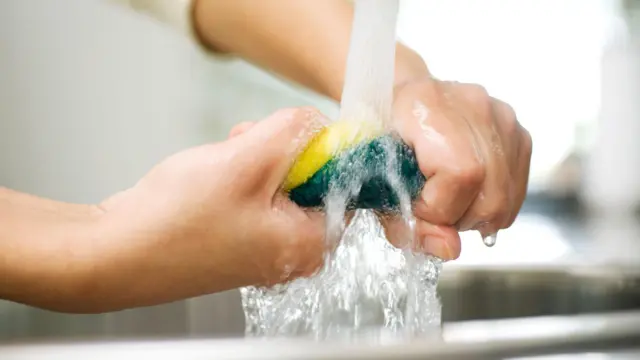 Foto de uma pessoa torcendo uma esponja de cozinha debaixo da torneira. 