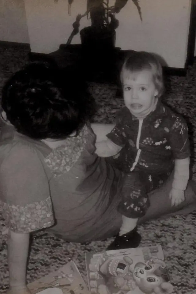 ஒரு கருப்பு வெள்ளை புகைப்படத்தில், ஒரு சிறிய குழந்தை (நினா) தரையில் தனது தாயின் மடியில் அமர்ந்திருக்கிறது. தாய் குனிந்து பார்ப்பதால் அவரது முகம் நமக்குத் தெரியவில்லை. அவர்களுக்கு முன்னால் தரையில் இரண்டு குழந்தைகளுக்கான புத்தகங்கள் கிடக்கின்றன.