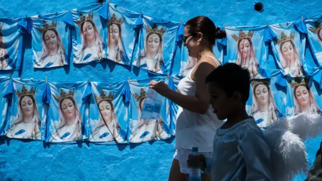 Uma mulher e uma criança vestida de anjo caminham em frente a um muro cheio de camisas da Imaculada Conceição, durante a festa em homenagem à santa católica no Recife, em 8 de dezembro de 2017