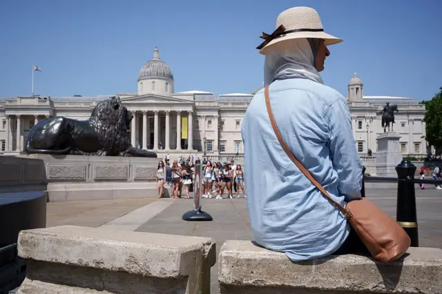 Mujer con camisa delgada y holgada de manga larga, protegida con un pañuelo y un sombrero, está sentada frente a la Galería Nacional de Londres.