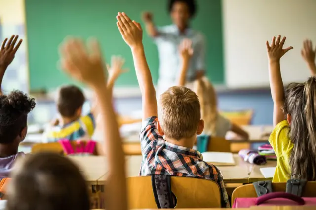 Crianças de costas sentadas em uma sala de aula com a mão para cima