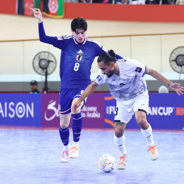 بازیکن افغانستان با لباس سفید پا به توپ، بازیکن جاپان با لباس نیلی و سفید به دنبال اوست که در پس زمینه آنها پرچم افغانستان نیز دیده می‌شود.