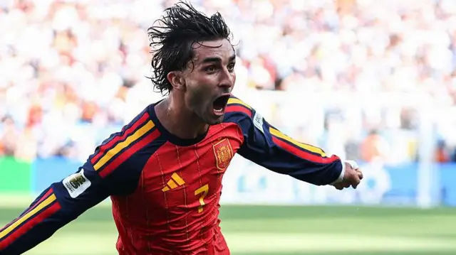 İspanyol futbolcu ellerini yana açarak koşuyor ve büyük bir sevinç yaşıyor. Üzerinde kızıl renkli İspanya forması var.