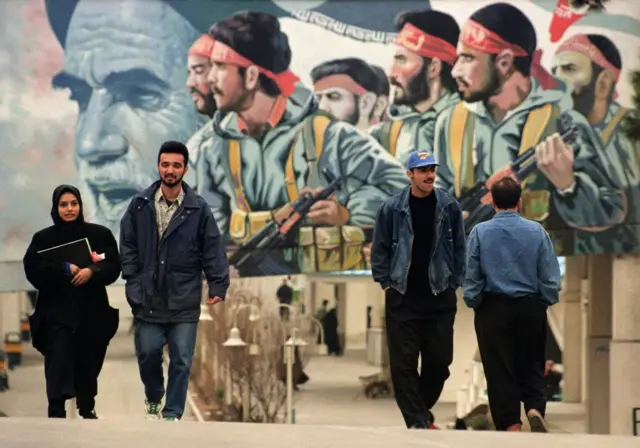شاب وفتاة يسيران في أحد شوارع طهران وخلفهما تظهر جدارية لعناصر من الحرس الثوري والمرشد الأعلى آية الله الخميني