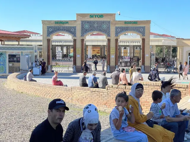 Siyob bozor kirishida odamlar bolalari bilan o'tiripti. Bolalar muzqaymoq yemoqda.