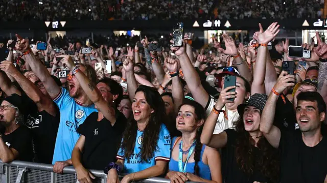 Fãs do Oasis no Principality Stadium. Um grande número de pessoas pode ser visto olhando para o palco. Muitos têm as mãos no ar e estão gravando o show em seus smartphones.
