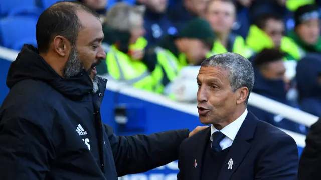 Nuno Espirito Santo, vêtu d'un imperméable à capuche bleu foncé, pose sa main gauche sur l'épaule de Chris Hughton tandis que les deux hommes discutent en bord de touche lors d'un match de football. Quatre personnes portant des gilets haute visibilité figurent parmi les personnes assises à l'arrière-plan, floues sur l'image.